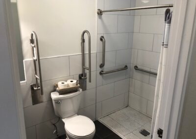 Accessible bathroom with a toilet, grab bars, and a roll-in shower. Two toilet paper rolls are on a basket above the toilet.