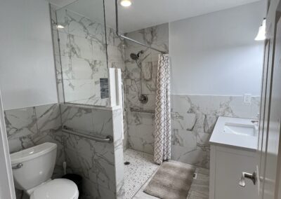 Modern bathroom with a walk-in shower, toilet, and sink. White and gray marble tiles cover the walls and floor. A patterned shower curtain and rugs add texture.