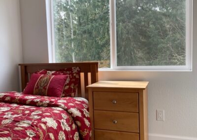 A bedroom corner features a wooden bed with red floral bedding and a matching nightstand. A large window shows a view of trees. The floor is wood-paneled.