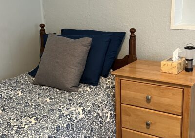 A neatly made bed with a blue floral bedspread and pillows next to a light wooden nightstand with a tissue box and a flashlight.