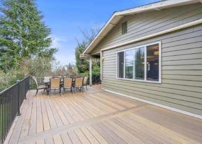 Wooden deck attached to a house with a dining table and chairs, surrounded by railing and overlooking trees under a clear blue sky.