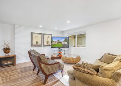 Cozy living room with a beige sofa, two armchairs, a wall-mounted TV, a small fireplace, and decorative artwork on a brick accent wall.