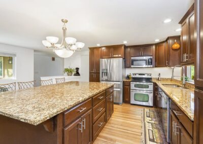 Modern kitchen with dark wood cabinets, stainless steel appliances, granite countertops, and a large island. Chandelier overhead and a wooden floor complete the space.